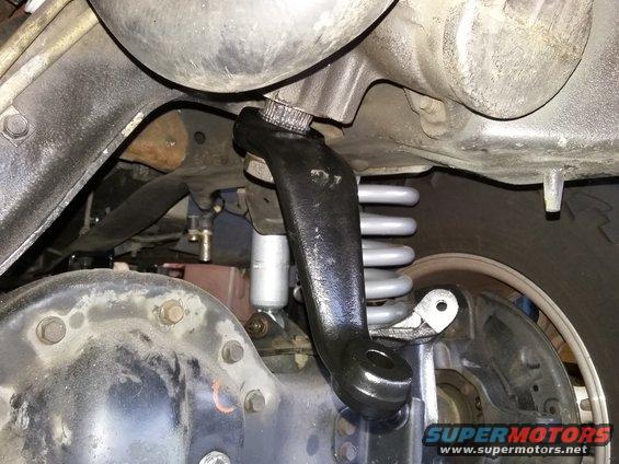 close-up of a vehicle's steering linkage component, possibly an idler arm or pitman arm, with suspension springs and axle visible