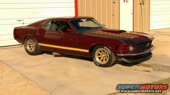 1970 Ford Mustang coupe with gold wheels and racing stripes parked in front of a garage