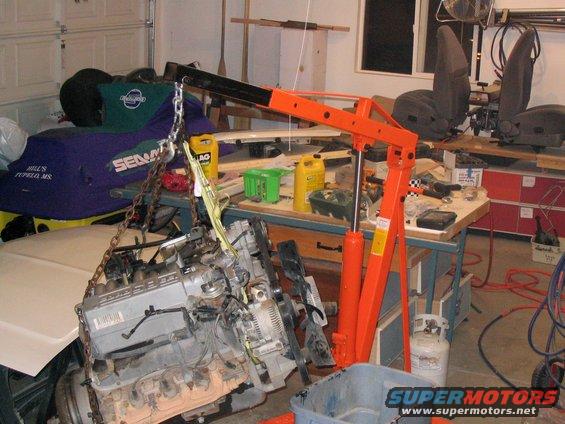 Engine suspended by an orange engine hoist in a garage. Other parts and tools are visible