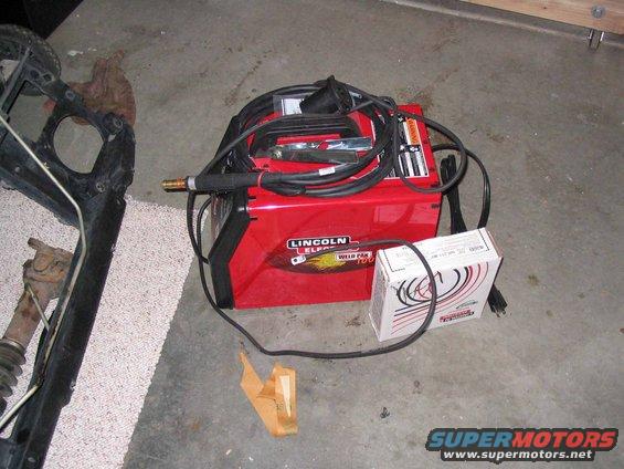 Red Lincoln Electric Weld Pak 100 welder with cables and a box of supplies on a concrete floor next to some vehicle parts