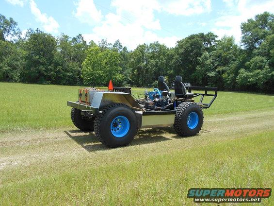 Custom off-road vehicle with exposed engine, two seats, and blue wheels, parked on a grassy field