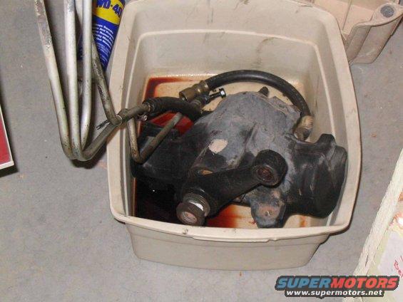 Power steering gearbox submerged in reddish fluid within a white plastic drain pan. Metal lines are attached