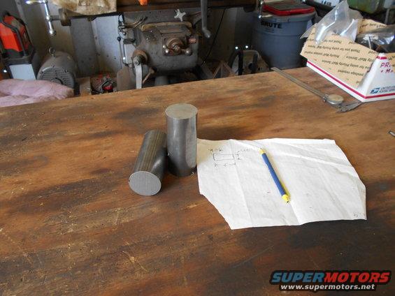 Two cylindrical metal stock pieces and a hand-drawn diagram with a pencil on a wooden workbench