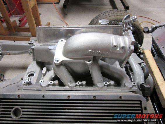 Close-up of an aluminum Edelbrock intake manifold on a V8 engine block, with a tire visible in the background