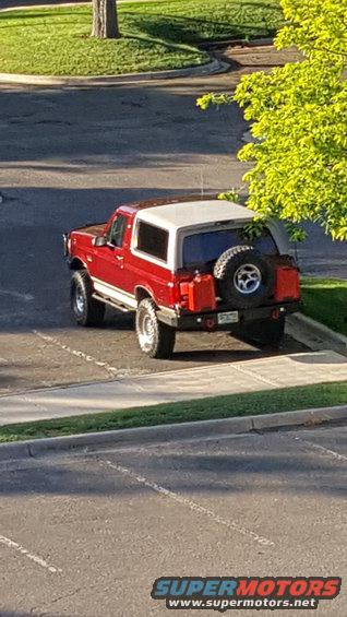 Red 1996 Ford Bronco with a white top, spare tire, and two red gas cans on the rear bumper, parked on asphalt