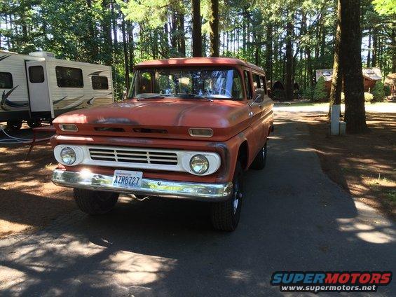 Orange Chevrolet Suburban with a white grille and chrome bumper, parked on asphalt near trees and a trailer
