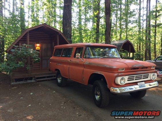 1962 Chevrolet Suburban, orange, parked in a wooded area next to a log cabin