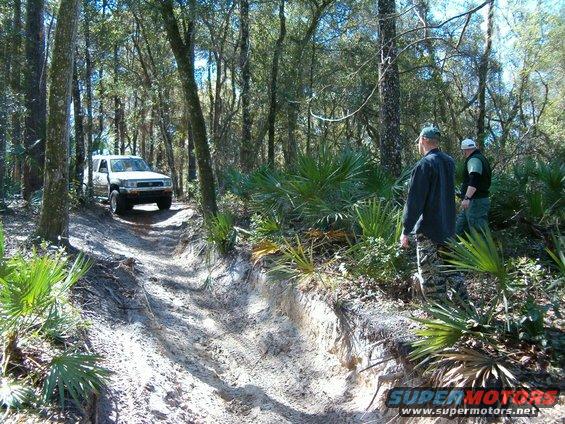 White Toyota 4Runner driving up a dirt trail in the woods. Two guys are standing on the side of the trail