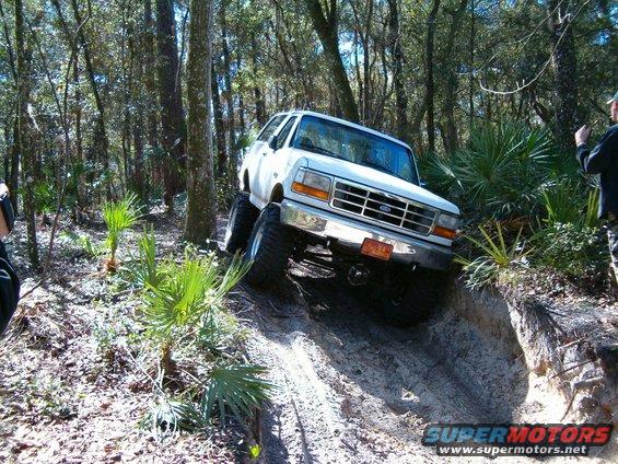 White Ford Bronco with large tires driving up a steep dirt incline in a wooded area