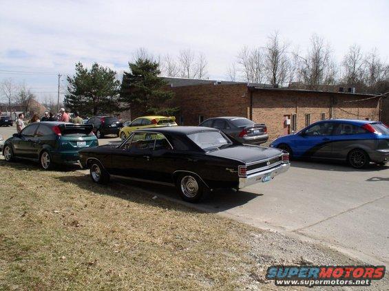 Several cars parked outside a brick building, including a black classic coupe, a green hatchback, and a blue and gray hatchback