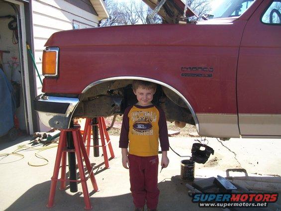 Red Ford Bronco lifted on jack stands, hood open, with a young boy in a yellow and maroon shirt standing in front