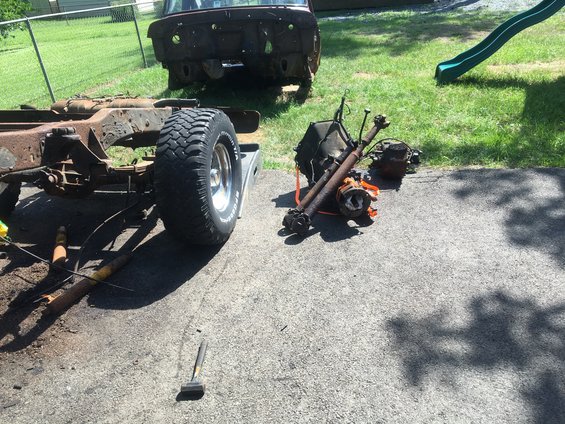 Vehicle chassis with one wheel and drivetrain parts like transmission and axles laid out on pavement next to a rusty vehicle body