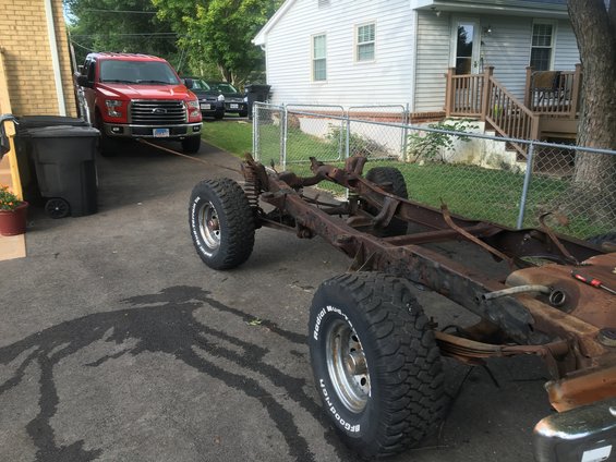 Rusty vehicle frame with BFGoodrich tires, pulled by a red Ford F-150 truck