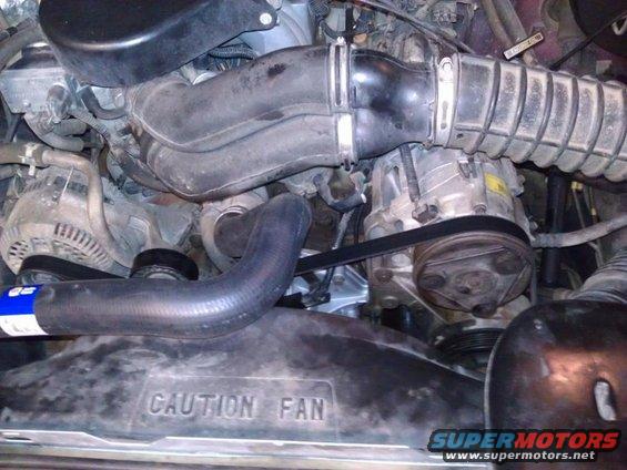 Engine bay with alternator, AC compressor, and fan shroud. Text on shroud reads 'CAUTION FAN'