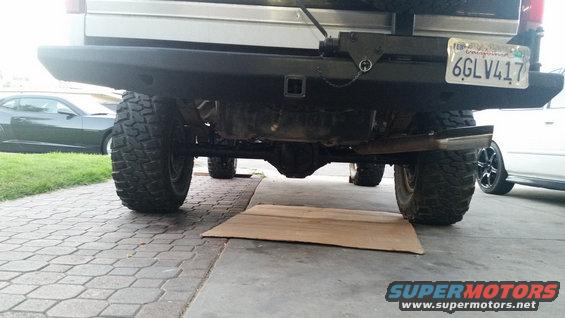 Rear view of a lifted SUV with large tires, aftermarket bumper, and trailer hitch. Another car is visible to the left