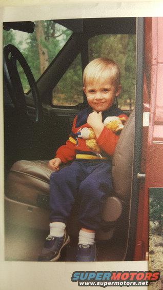 Child in red and blue outfit sits in a vehicle's passenger seat, holding a stuffed animal
