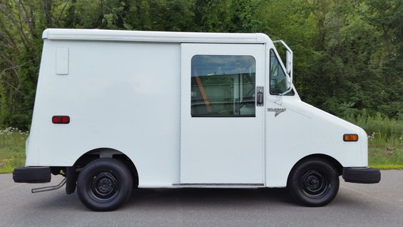 White Grumman LLV postal truck, side view. It has a sliding door and a window