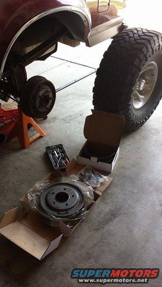 Maroon Ford Bronco rear wheel removed, supported by jack stands, with new brake parts on the floor