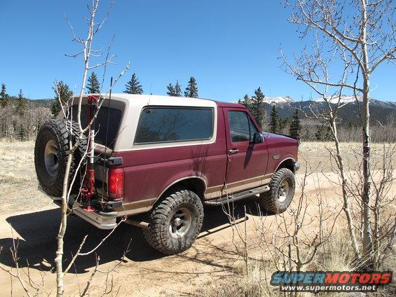Maroon 1996 Ford Bronco parked on a dirt road with a spare tire mounted on the rear swing-away carrier