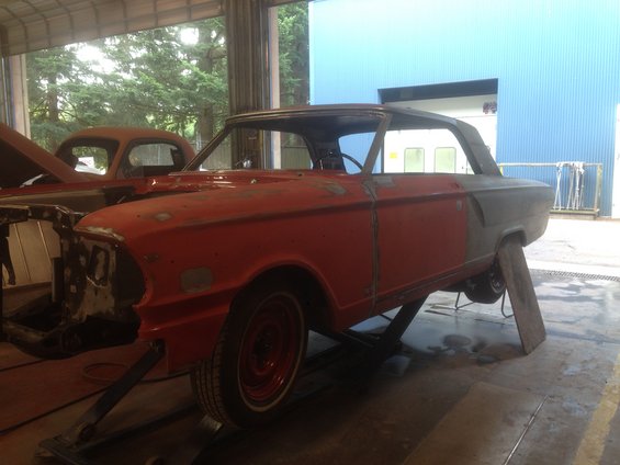 Red 1964 Ford Fairlane coupe body on a lift, undergoing repairs, with another car visible behind