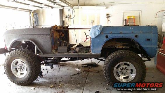 Ford Bronco body, stripped down and on a lift in a garage. It has large off-road tires and appears to be undergoing restoration
