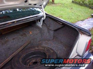 Car trunk interior with spare tire and wood plank. Gray carpet lining. Watermark "SUPERMOTORS.NET"