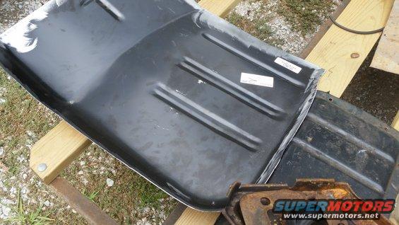Close-up of a black metal vehicle floor pan with raised ribs, resting on wooden beams. Two white stickers are visible on the surface