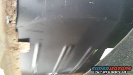 Close-up of a vehicle's undercarriage, showing some rust on the left and a dark panel with ribbed reinforcement