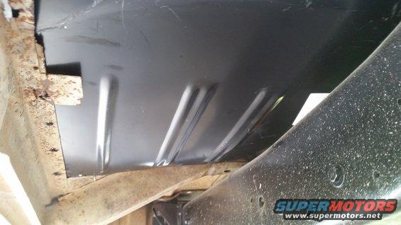 Undercarriage of a vehicle showing a black metal floor panel with ribbed reinforcement and rusty frame components
