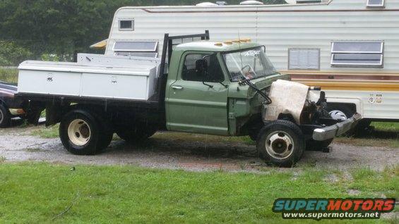 Green flatbed truck with a white toolbox on the bed, parked next to a camper. Front end is partially disassembled