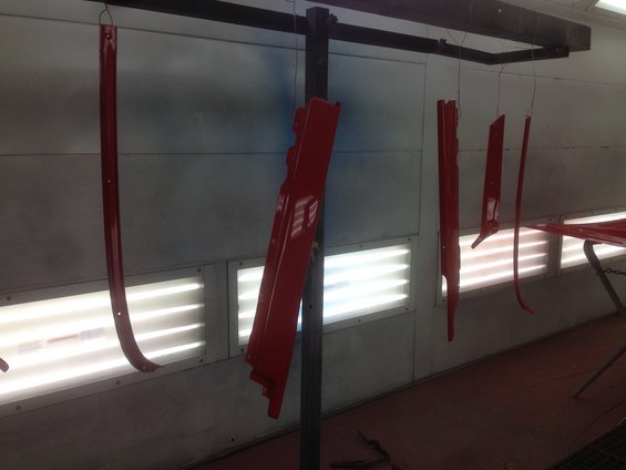 Red car body panels hanging in a paint booth, illuminated by fluorescent lights