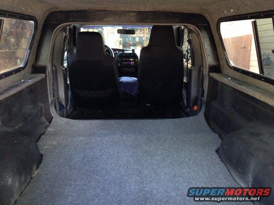 Interior view of a pickup truck bed with carpet, showing the back of the front seats and a camper shell