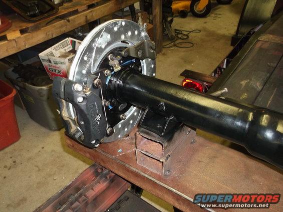 Rear axle assembly with a drilled disc brake rotor and black caliper, supported by metal blocks on a workbench