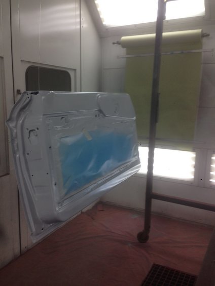 Car door covered in blue plastic tape inside a white paint booth