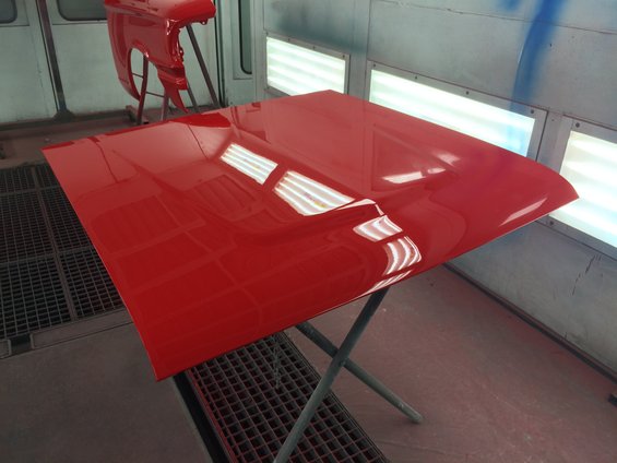 Red car hood and fender on stands inside a paint booth with lights reflecting on the hood