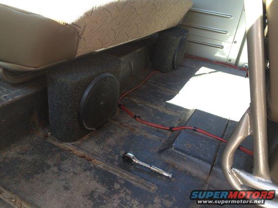 Vehicle interior with two subwoofers under a tan seat, red wiring, and a socket wrench on the floor