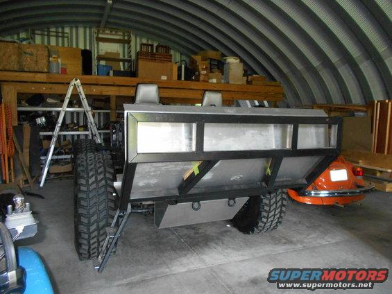 Trailer chassis with large off-road tires, metal bed frame, and two headrests, parked in a garage next to an orange car