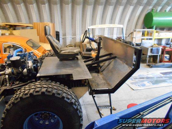 Custom off-road vehicle chassis with large tires, engine, and basic seating. Orange VW Beetle visible in background