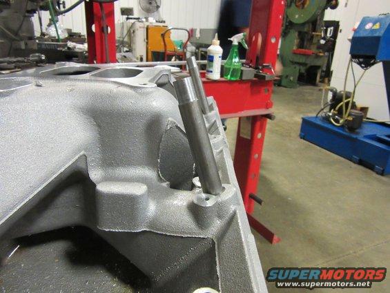 Close-up of an engine intake manifold with two metal rods inserted into holes. Shop equipment is visible in the background