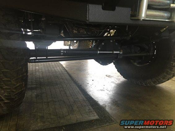 Undercarriage view showing the rear axle, steering linkage, and large tires of a vehicle. Supermotors.net logo visible