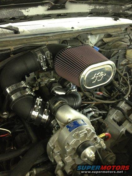 Engine bay with K&N air filter, black intake tubing, and a silver supercharger. Some wiring and hoses are visible