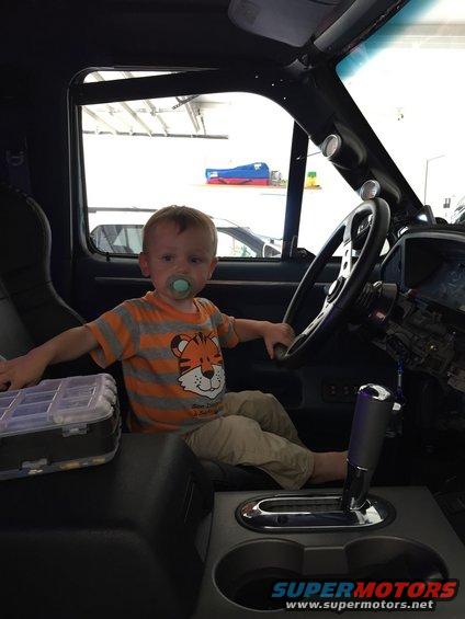 Kid with pacifier in driver's seat, holding steering wheel. Toolbox on console