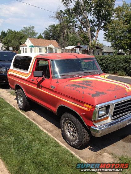 Red 1978 Ford Bronco parked on a street, showing its side and front quarter. It has racing stripes on the hood and a white camper shell