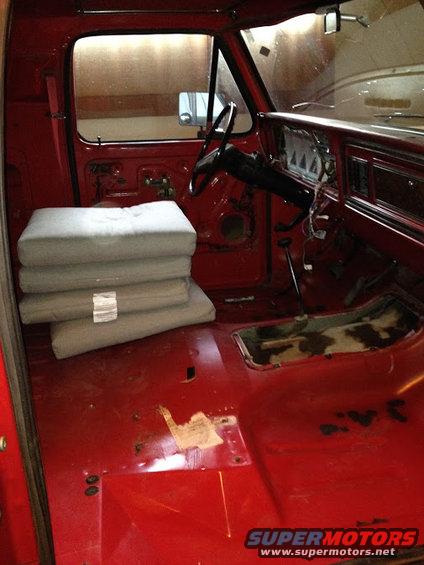 Interior of a red truck cab, showing the dashboard, steering wheel, and seats removed. Stacked gray padding is on the passenger side