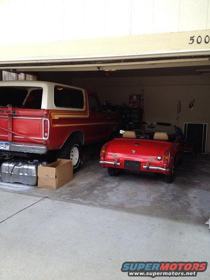 Red 1978 Ford Bronco with white top and side stripes, parked next to a red convertible in a garage