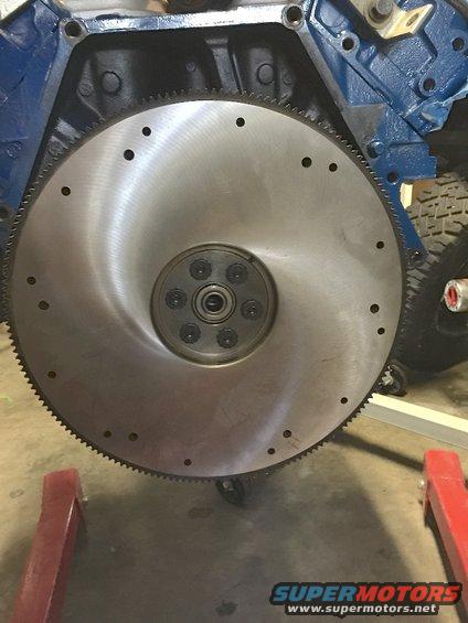 Metal flywheel with bolted center and gear teeth on edge, attached to blue engine block