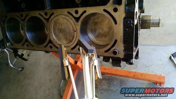 Engine block with three cylinders visible, pistons in two, and a honing tool in place. The block is on an orange engine stand