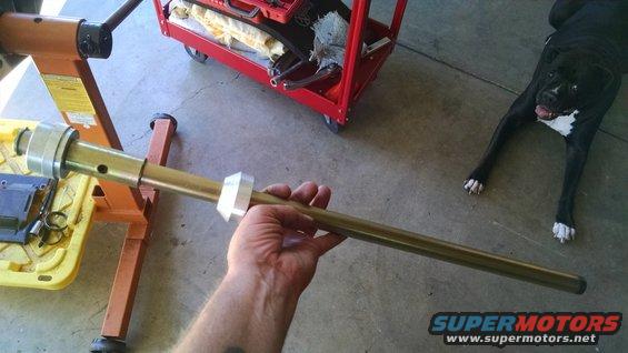 Hand holding a brass-colored steering column component with silver collars. Red tool cart and black dog in background