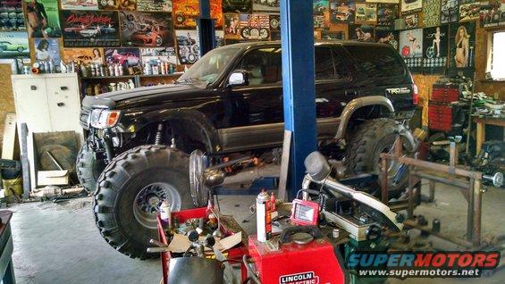 Black Toyota 4Runner on lift with huge tires, in a garage with posters on the wall