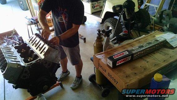 Man working on engine block with COMP Cams camshaft box nearby. A golf cart is visible in the background